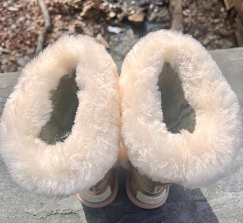 May include: A pair of beige winter boots with a soft, fluffy, cream-colored lining. The boots have a light pink sole and a metallic gold exterior. The boots are designed to keep feet warm in cold weather.