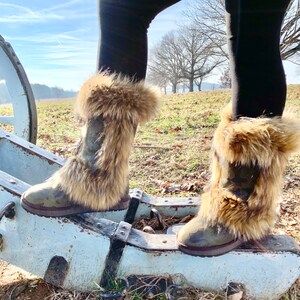 Peut inclure: Une paire de bottes marron &agrave; fourrure synth&eacute;tique avec un motif camouflage. Les bottes sont port&eacute;es par une personne debout sur une structure en bois.