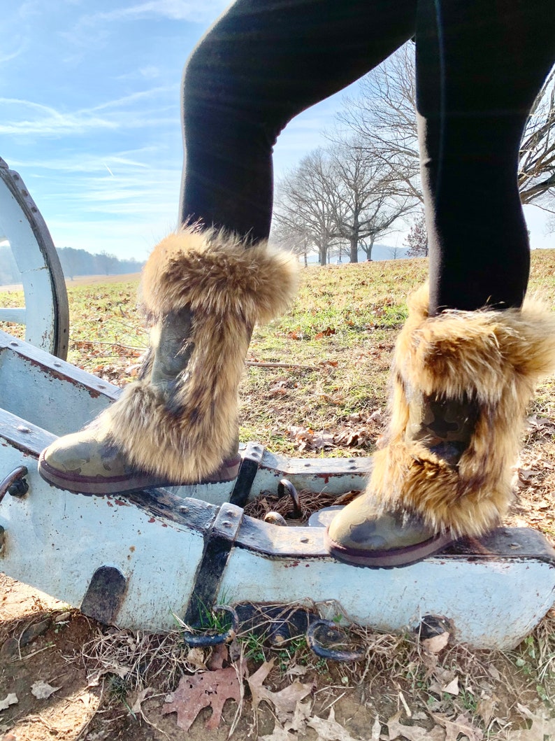 Peut inclure: Une paire de bottes marron &agrave; fourrure synth&eacute;tique avec un motif camouflage. Les bottes sont port&eacute;es par une personne debout sur une structure en bois.