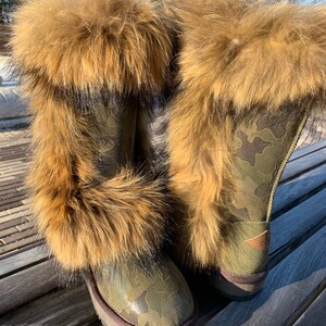 Peut inclure: Une paire de bottes de camouflage vert avec une bordure en fourrure brune. Les bottes sont pos&eacute;es sur un banc en bois.