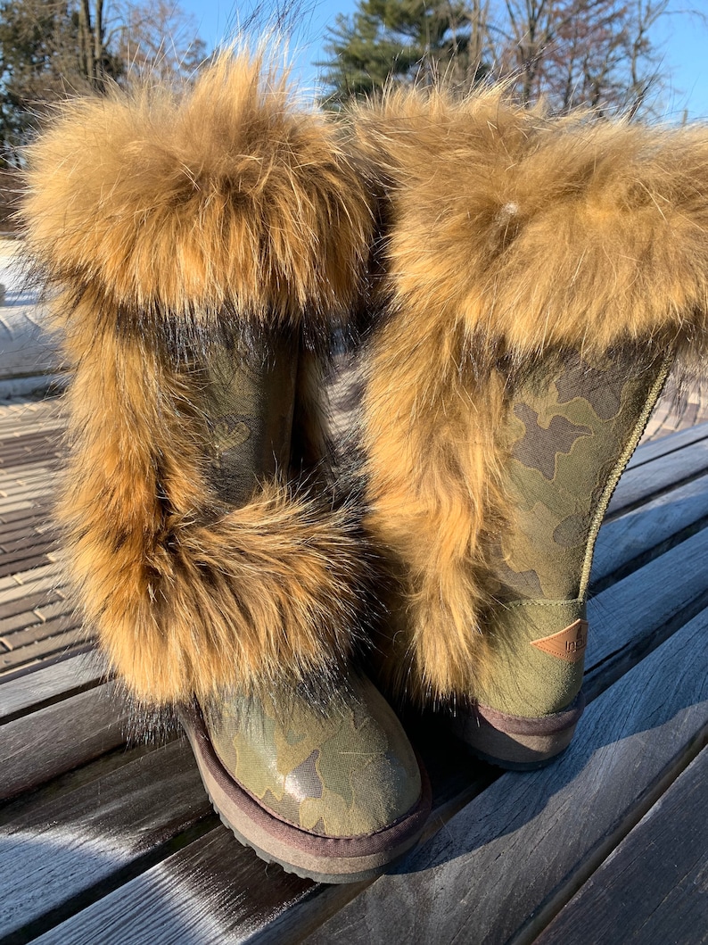 Peut inclure: Une paire de bottes de camouflage vert avec une bordure en fourrure brune. Les bottes sont pos&eacute;es sur un banc en bois.