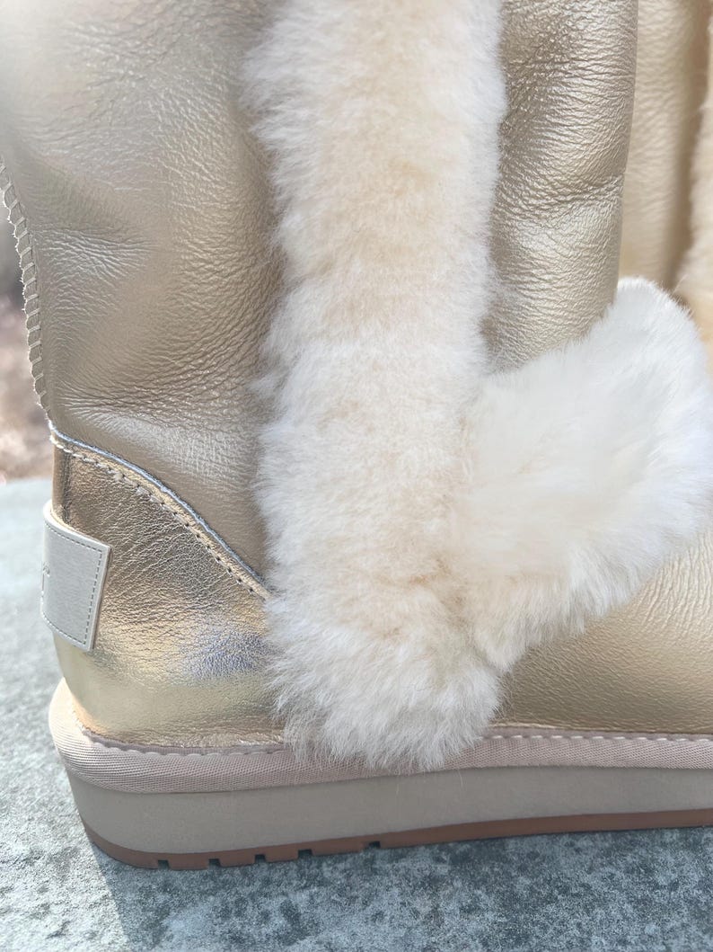 May include: Close-up of a gold-colored boot with cream-colored fur trim. The boot has a metallic sheen and a thick, beige sole. The fur trim is fluffy and contrasts with the smooth leather-like material of the boot.