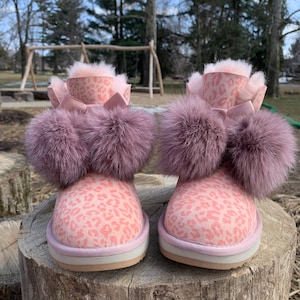 Puede incluir: Botas de piel sintética rosa con estampado de leopardo y dos pompones grandes en cada bota.
