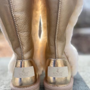 May include: A pair of gold-colored boots with a light tan interior and a light pink sole. The boots have a metallic sheen and a label on the back that reads "Inoli Luxury Reinterpreted". The boots are made of leather and sheepskin.