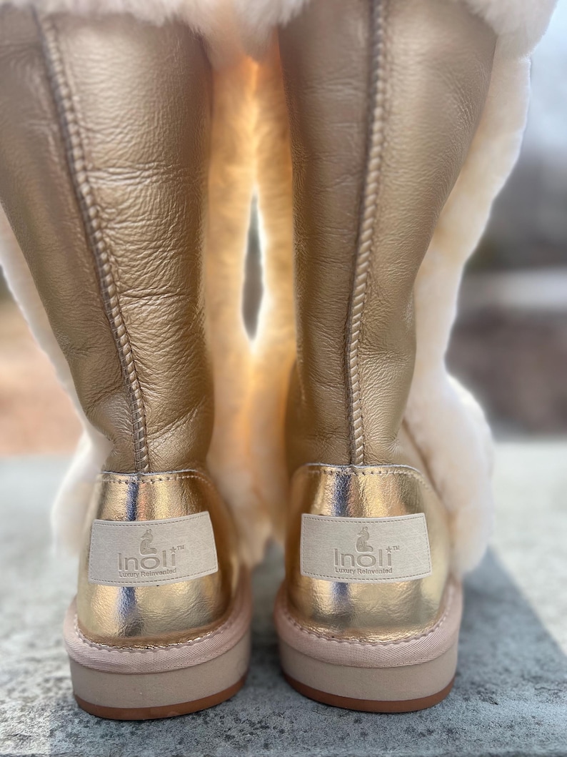May include: A pair of gold-colored boots with a light tan interior and a light pink sole. The boots have a metallic sheen and a label on the back that reads "Inoli Luxury Reinterpreted". The boots are made of leather and sheepskin.