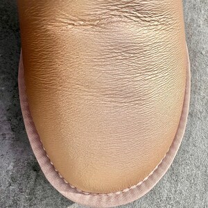 May include: Close-up of a tan leather shoe with a rounded toe. The shoe has a textured surface and is trimmed with a light pink fabric. The stitching is visible along the edge of the fabric.