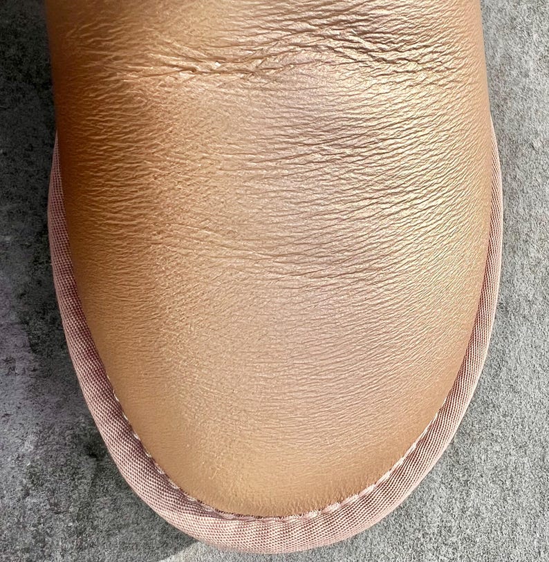 May include: Close-up of a tan leather shoe with a rounded toe. The shoe has a textured surface and is trimmed with a light pink fabric. The stitching is visible along the edge of the fabric.