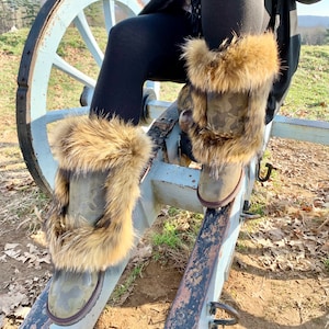Peut inclure: Une paire de bottes de camouflage brun et vert avec une bordure en fausse fourrure. Les bottes sont port&eacute;es par une personne assise sur une roue de canon en bois.