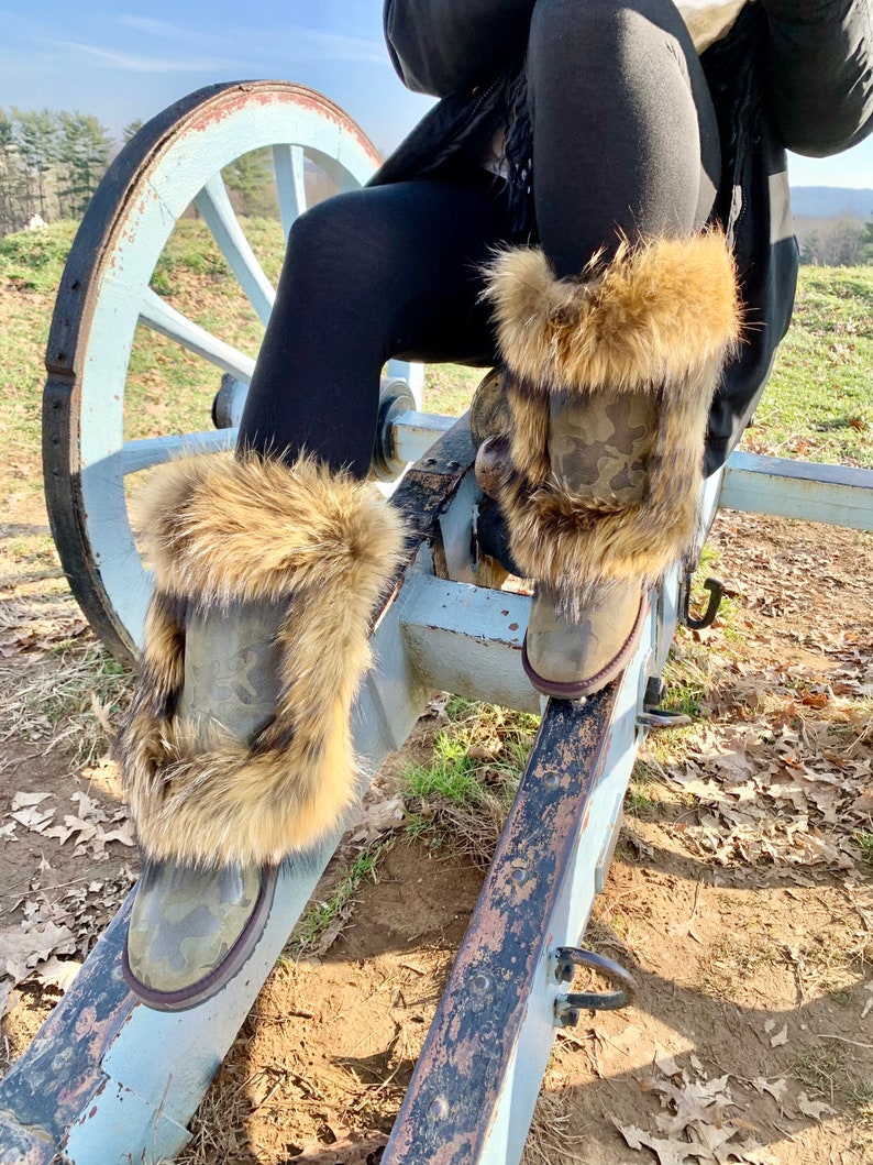 Peut inclure: Une paire de bottes de camouflage brun et vert avec une bordure en fausse fourrure. Les bottes sont port&eacute;es par une personne assise sur une roue de canon en bois.