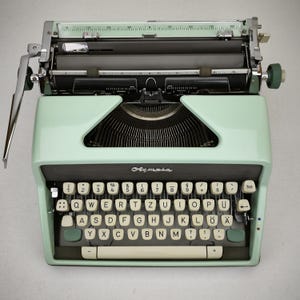 May include: A vintage mint green Olympia typewriter with a black keyboard and white keys. The keys are arranged in a QWERTZ layout. The typewriter has a chrome carriage return lever and the word "Olympia" is printed on the front.