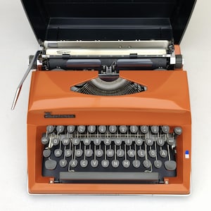 May include: A vintage orange Triumph Contessa portable typewriter with a QWERTZ keyboard layout, black circular keys, a metal carriage, and a black top cover. The brand name Triumph is printed on the carriage, and Contessa is on the front panel.