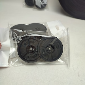 Peut inclure: Plusieurs bobines de ruban d'encre noires pour machine à écrire sont visibles dans un emballage plastique transparent. Les bobines ont un design circulaire avec un trou central et des rayons. D'autres bobines sont visibles en arrière-plan.