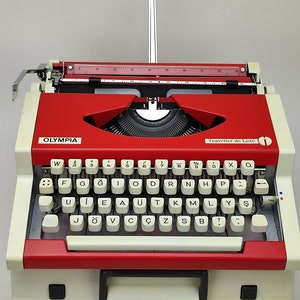 May include: A red and white Olympia Traveller de Luxe typewriter with a black keyboard and white keys. The keys have letters in both English and German.