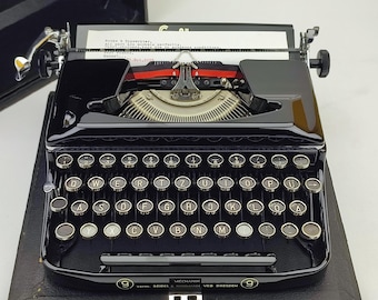 May include: A black vintage typewriter with a silver carriage return lever and a black case. The typewriter has a German keyboard layout with the text "MECHANIK 9 verm. SEIDEL & NAUHANN VEB DRESDEN 9" on the front.
