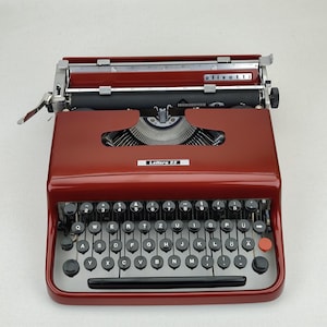 May include: A dark red Olivetti Lettera 22 portable typewriter featuring a QWERTZ keyboard layout with black circular keys, a silver metal carriage, and a prominent red key on the right side. The brand name and model are printed on the front casing.