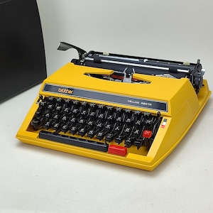 May include: A yellow Brother Deluxe 880TR typewriter with black keys and a red key. The typewriter has the brand name "brother" and "DELUXE 880TR" printed on it. The typewriter is in a light-colored room.