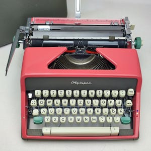 Può includere: Una macchina da scrivere rossa con una tastiera nera e grigia. I tasti hanno lettere bianche e accenti verdi. La macchina da scrivere ha una leva di ritorno a capo nera e un tasto di tabulazione nero.