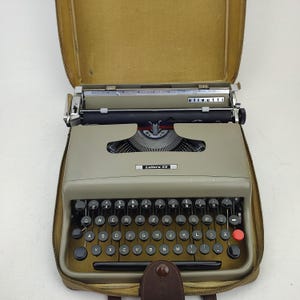 May include: A classic Olivetti Lettera 22 typewriter in a tan case. The typewriter is a pale grey with black keys and a red return key. The word "olivetti" is visible on the top, and "Lettera 22" is printed on the front.