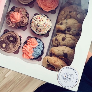May include: A white box filled with a variety of cupcakes and chocolate chip cookies. The cupcakes have different colored frostings and toppings. The cookies are stacked on top of each other. The box has a sticker on it that says "Bake Eat Repeat" and a website address.