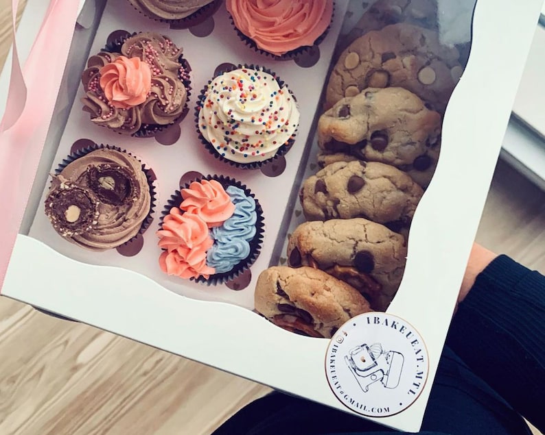 May include: A white box filled with a variety of cupcakes and chocolate chip cookies. The cupcakes have different colored frostings and toppings. The cookies are stacked on top of each other. The box has a sticker on it that says "Bake Eat Repeat" and a website address.