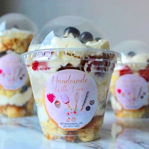 May include: Three clear plastic cups filled with a layered dessert. The dessert includes cake, whipped cream, fresh fruit, and blueberries. The cups have a pink and white sticker that says "Handmade with Love" and includes a logo and contact information.