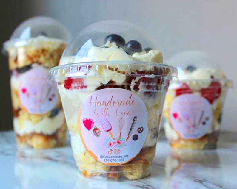 May include: Three clear plastic cups filled with a layered dessert. The dessert includes cake, whipped cream, fresh fruit, and blueberries. The cups have a pink and white sticker that says "Handmade with Love" and includes a logo and contact information.