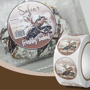 May include: A roll of round stickers with a brown background and a picture of a chocolate pastry with powdered sugar on top. The sticker says "Sofia's freshly baked" and lists the ingredients: flour, cocoa, eggs, cinnamon, and ginger. The sticker also says "Made in USA".