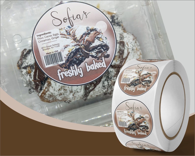 May include: A roll of round stickers with a brown background and a picture of a chocolate pastry with powdered sugar on top. The sticker says "Sofia's freshly baked" and lists the ingredients: flour, cocoa, eggs, cinnamon, and ginger. The sticker also says "Made in USA".