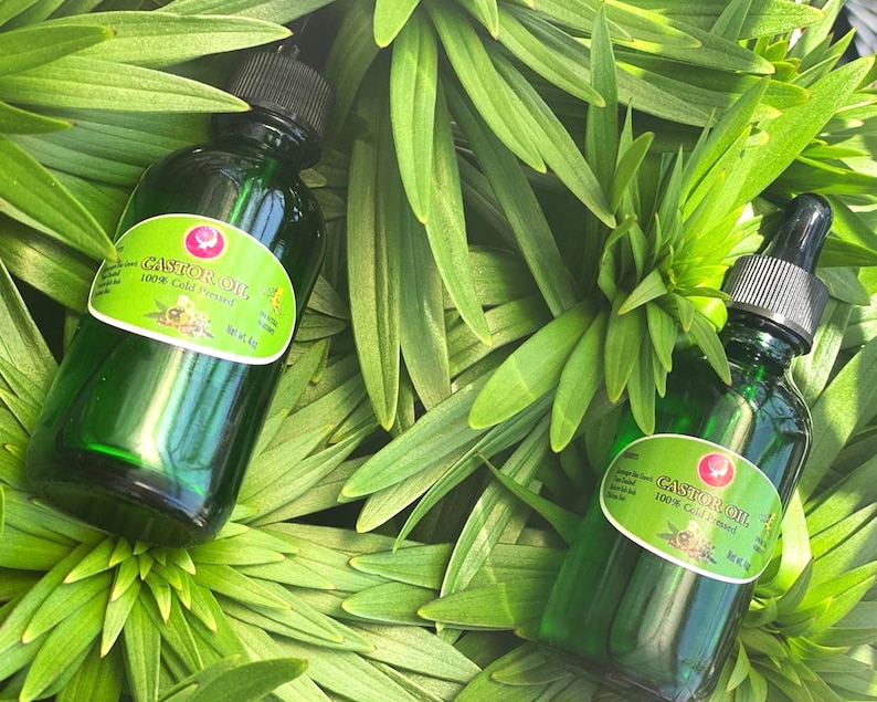 May include: Two green glass bottles of castor oil, labeled "Castor Oil 100% Cold Pressed" with a white label and a gold logo. The bottles are sitting on a bed of green leaves.