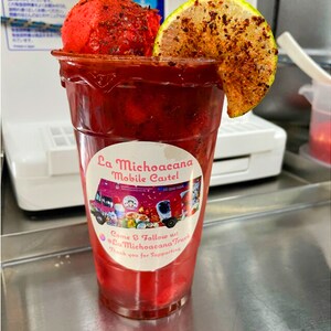 May include: A red, frozen drink with a lime wedge and a sprinkle of chili powder in a clear plastic cup. The cup has a sticker that says "La Michoacana Mobile Castel" and "Come & Follow us: @LaMichoacanaTruck".
