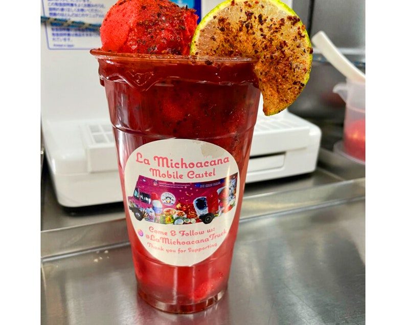 May include: A red, frozen drink with a lime wedge and a sprinkle of chili powder in a clear plastic cup. The cup has a sticker that says "La Michoacana Mobile Castel" and "Come & Follow us: @LaMichoacanaTruck".
