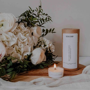 Puede incluir: Una vela blanca con un tubo de cartón marrón detrás. El tubo tiene el texto "Wild Child" y "Kit Découverte". La vela está encendida y hay un ramo de flores blancas y rosas en el fondo.