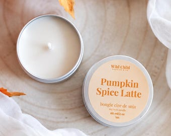 May include: A round silver metal tin containing a white soy wax candle with a single wick. The lid features a beige label with the text Wild Child, Pumpkin Spice Latte, bougie cire de soja, soy wax candle, 60 ml/2 oz, and 15h printed in brown.