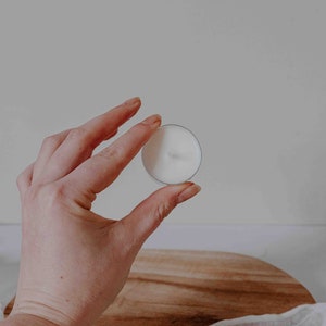 Może przedstawiać: Biała świeczka typu tealight w przezroczystym szklanym pojemniku. Świeczka jest trzymana w dłoni.