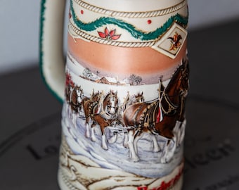 May include: A white ceramic beer stein featuring a winter scene with a team of horses pulling a wagon. The stein has green and red trim and the words "Budweiser" and "Holiday" are visible.