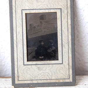 May include: Vintage black and white photograph in a decorative frame. The frame has a gray border and a patterned interior. The photograph depicts a large ship and two people in dark clothing. The image is aged and has a nostalgic feel.