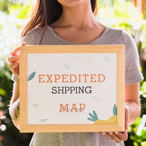 May include: A person holding a framed sign with the text "EXPEDITED SHIPPING MAP" in orange and black lettering. The sign has a white background with a colorful design of leaves, swirls, and a paper airplane.