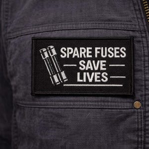 May include: Black rectangular patch with the text "SPARE FUSES SAVE LIVES" in white, along with a white illustration of two fuses. The patch is sewn onto a dark gray jacket.