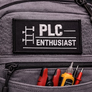 Könnte beinhalten: Grauer Rucksack mit einem schwarzen Aufnäher mit der weißen Aufschrift "PLC ENTHUSIAST". Die Vordertasche enthält rote und gelbe Werkzeuge. Die Tasche hat einen schwarzen Reißverschluss und eine schwarze Kordel.