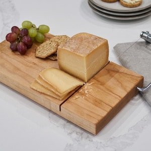 May include: A wooden cheese board with a block of cheese, sliced cheese, crackers, and grapes. A cheese slicer and a linen napkin are also present. The board is made of light-colored wood.