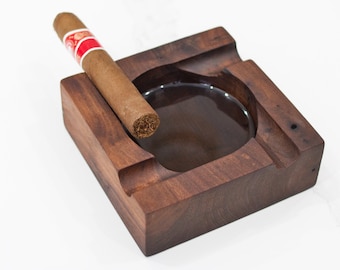 May include: A dark brown wooden cigar ashtray with a round, recessed area for ashes. A brown cigar is resting on the side of the ashtray.