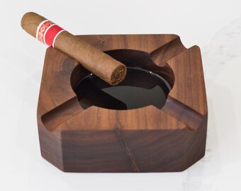 May include: A dark brown wooden cigar ashtray with a square shape and a deep well. A brown cigar is resting on the edge of the ashtray.