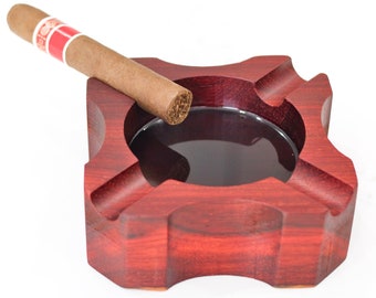 May include: A red wooden cigar ashtray with a cigar resting on the edge. The ashtray has a deep well for ash and a separate groove for resting the cigar.