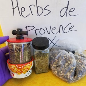 May include: Three containers of herbs and spices. A red and white ceramic grinder with a metal top filled with dried herbs. A clear glass jar with a black lid filled with dried herbs. A clear plastic bag filled with black, white, and brown peppercorns. The label on the white box says "Herbs de Provence".