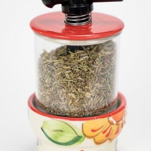 May include: A ceramic herb grinder with a red lid and a black handle. The grinder is filled with dried herbs and has a floral design on the bottom.