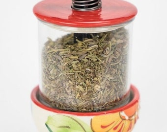 May include: A ceramic herb grinder with a red lid and a black handle. The grinder is filled with dried herbs and has a floral design on the bottom.