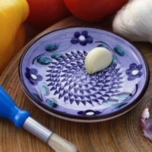 May include: A ceramic garlic grater with a blue handle and a blue and purple floral design. A clove of garlic sits on the grater. Other garlic bulbs, a yellow bell pepper, and a tomato are in the background.