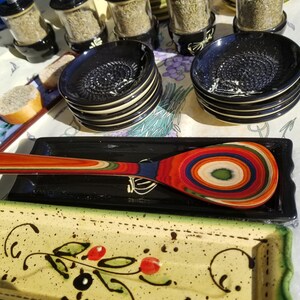 May include: A set of black and white ceramic garlic grater dishes with a wooden spoon with colorful stripes. The dishes are stacked on top of each other and the spoon is lying on top of a black and white dish. The dishes have a decorative design on the bottom.