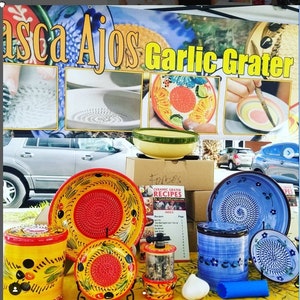 May include: A collection of colorful ceramic garlic graters, some with a yellow and red design, others with a blue and white design. The graters are displayed on a table with a sign that reads "Asca Ajos Garlic Grater".