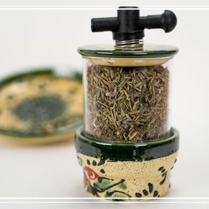 May include: A ceramic herb grinder with a green and yellow floral design. The grinder has a clear glass jar filled with dried herbs and a black plastic handle on top.